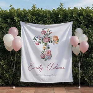 May include: White fabric backdrop with a floral cross design, featuring pink and peach roses, and other flowers. The text "Emily Adams, Christening Day, June 12, 2026" is printed on the backdrop. Pink and white balloons are on either side.