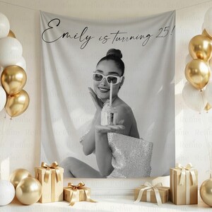 May include: Black and white banner with the text "Emily is turning 25!" A woman in sunglasses holds a cupcake with a lit candle. Gold and white balloons and wrapped gifts with gold ribbons are in the background.