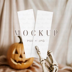 Könnte beinhalten: Ein Halloween-Themen-Mockup mit zwei weißen rechteckigen Karten, die von einer Skeletthand gehalten werden. Im Hintergrund befindet sich ein geschnitzter orangefarbener Kürbis mit einem lächelnden Gesicht. Das Wort "MOCKUP" wird über "PSD + JPG" angezeigt.