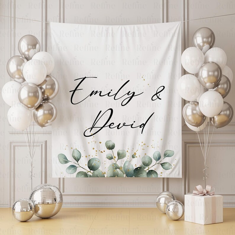 May include: A white backdrop with the names "Emily & Devid" in elegant script, adorned with eucalyptus and gold accents. Silver and white balloons flank the backdrop, with disco balls and a gift box completing the festive scene.