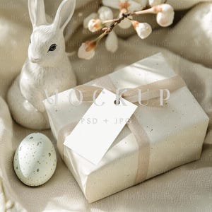 May include: A white gift box with a beige ribbon and a white gift tag that says "MOCKUP PSD + JPG". The box is sitting on a beige fabric with a white ceramic bunny and a speckled blue Easter egg.