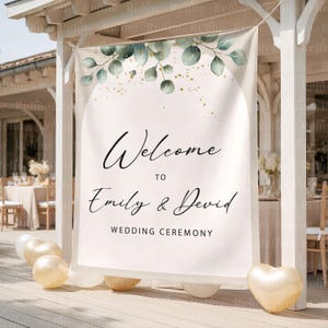 Op de afbeelding: Een witte trouwceremoniebanner met de tekst "Welcome to Emily & David" en "Wedding Ceremony" in zwart schrift. De banner is versierd met groene eucalyptusbladeren en gouden accenten. Gouden ballonnen staan aan de basis.
