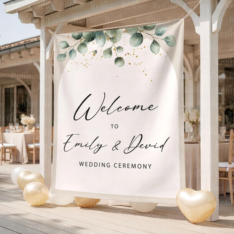 Op de afbeelding: Een witte trouwceremoniebanner met de tekst "Welcome to Emily & David" en "Wedding Ceremony" in zwart schrift. De banner is versierd met groene eucalyptusbladeren en gouden accenten. Gouden ballonnen staan aan de basis.