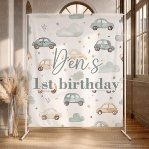 May include: A white backdrop featuring watercolor illustrations of cars, clouds, and stars. The text "Den's 1st birthday" is displayed in a script font. The backdrop is supported by a white metal frame, suitable for a birthday party.