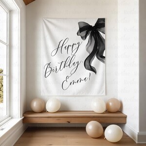 K&ouml;nnte beinhalten: Ein wei&szlig;es Banner mit dem Text "Happy Birthday Emma!" in eleganter Schrift, akzentuiert durch eine gro&szlig;e schwarze Schleife. Mehrere beige Luftballons sind um eine Holzbank herum verstreut und schaffen eine festliche Geburtstagsatmosph&auml;re.