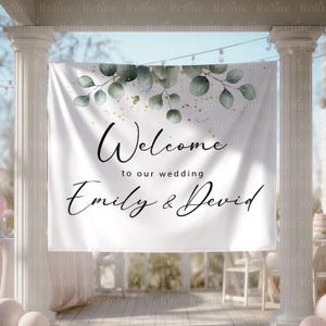 May include: A white banner with the text "Welcome to our wedding Emily & David" in elegant script. The banner is decorated with green eucalyptus leaves and gold accents, perfect for a wedding ceremony.