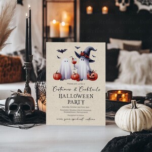 Op de afbeelding: Halloween-feestuitnodiging met aquarelgeesten, pompoenen en vleermuizen. De uitnodigingstekst luidt "Costumes & Cocktails" en bevat evenementdetails. Een zwarte schedelkaarsenhouder en een witte pompoen zijn ook zichtbaar.
