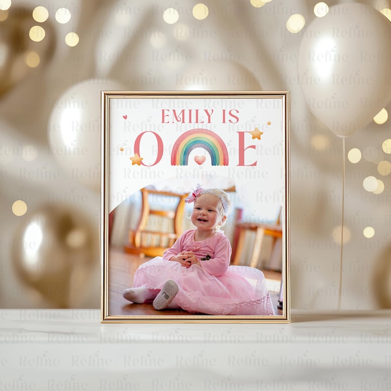 May include: A gold-framed photo with a white background and a rainbow with a pink heart in the center. The text "Emily is One" is written in pink letters with gold stars. A photo of a young girl in a pink dress is in the bottom half of the frame.