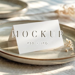 May include: A white folded card with the word "MOCKUP" printed on it, resting on a ceramic plate. The plate has a light brown rim and is placed on a textured surface. The text "PSD + JPG" is also visible on the card.
