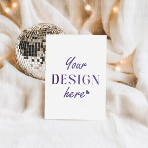 Puede incluir: Una tarjeta blanca con el texto "Your DESIGN here" en escritura p&uacute;rpura. Una bola de discoteca plateada est&aacute; a la izquierda, sobre una tela blanca texturizada. Peque&ntilde;as luces son visibles en el fondo, creando un ambiente festivo.