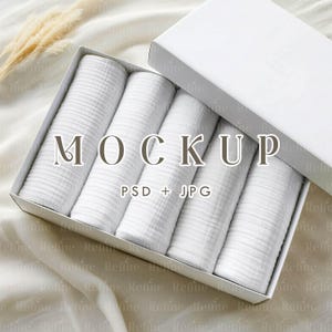 May include: A white box with a partially open lid, containing six rolled white fabric items. The word "MOCKUP" is displayed in the center, with "PSD + JPG" below it. The box is on a white surface.
