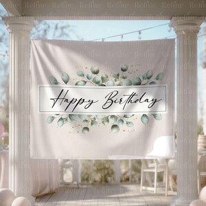 May include: A beige banner with the words "Happy Birthday" in elegant script. The banner is decorated with green eucalyptus leaves and gold flecks. It is hanging between white pillars, suggesting an outdoor party setting.