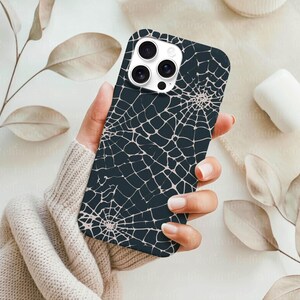 May include: A dark navy blue phone case featuring a spiderweb design in a light beige color. The case has a glossy finish and is designed to fit a smartphone. The case is held in a person's hand.
