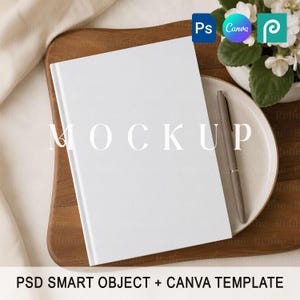 May include: A blank white notebook with a smooth cover positioned on a round ceramic plate next to a metallic pen. The notebook is placed on a wooden board, with a small cluster of white flowers visible in the upper right corner of the frame.