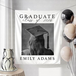 May include: A white graduation banner with the text "GRADUATE class of 2026" and "EMILY ADAMS". The banner features a black and white photo of a person wearing a graduation cap. Balloons and a cake are also visible.