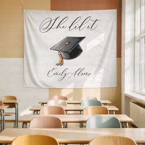 May include: White graduation banner with the text "She did it" in elegant script, a black graduation cap illustration with a gold tassel, and the name "Emily Alums" below. The banner is hanging in a classroom.