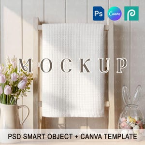 Op de afbeelding: Een witte wafelweefsel handdoek hangt aan een houten rek, met het woord "MOCKUP" in het midden. Aan weerszijden staan een vaas met roze en witte bloemen en een glazen konijnenpot gevuld met snoep. De tekst "PSD SMART OBJECT + CANVA TEMPLATE" is inbegrepen.