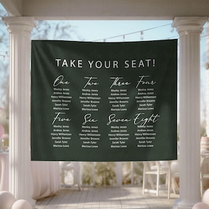 May include: A dark green wedding seating chart banner with white calligraphy text that reads "TAKE YOUR SEAT!" and lists names under numbers one through eight. The banner is hanging outdoors.
