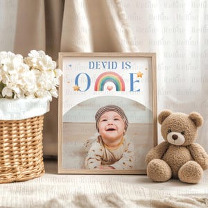 May include: A framed photo with a rainbow and the text "DEVID IS ONE" in blue letters. A baby boy is smiling and looking at the camera. The photo is on a white surface with a brown teddy bear.
