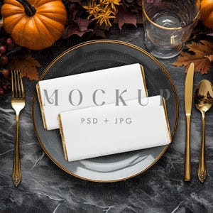 May include: A mock-up of a table setting with a dark gray plate with a gold rim, two white rectangular objects with gold edges, and gold silverware. A pumpkin and fall foliage are in the background.