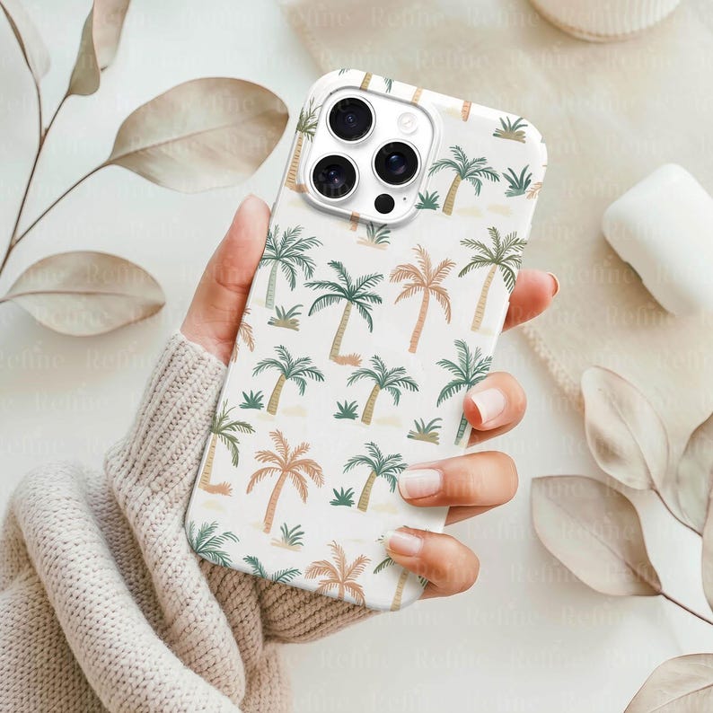 May include: A white phone case featuring a repeating pattern of palm trees in shades of green, brown, and tan. The case is held in a person's hand, showcasing the design's tropical theme. The phone case has a camera cutout.