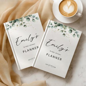 May include: Two white bridal shower planners with the name "Emily's" in elegant script. The planners have a botanical design with green leaves and gold accents. The text "bridal shower PLANNER" is below the name, with the date "SAT 11" on one and "SAT 11 FEB" on the other.