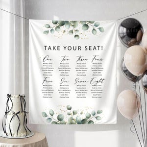 May include: A white wedding seating chart with eucalyptus and gold accents. The chart is divided into eight sections, each listing names. The text "TAKE YOUR SEAT!" is at the top. A two-tiered cake and balloons are in the image.