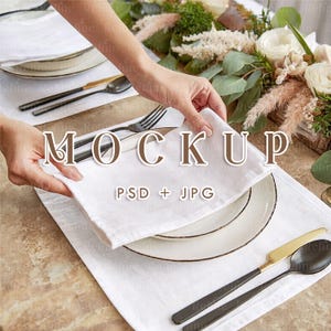 Puede incluir: Una servilleta de lino blanco se coloca sobre un plato blanco con borde negro. La mesa está puesta con cubiertos negros y dorados, un mantel individual blanco y un arreglo floral. La palabra "MOCKUP" se muestra en la imagen.