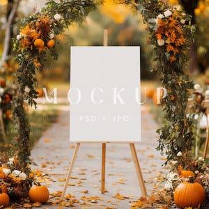 Pode incluir: Um cavalete de madeira com uma placa branca em branco, adornada com folhagem de outono e abóboras. A placa diz "MOCKUP PSD + JPG". A cena é ambientada ao ar livre com um fundo desfocado de cores de outono.