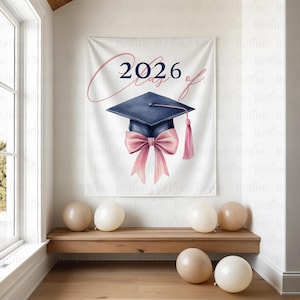 K&ouml;nnte beinhalten: Ein wei&szlig;es Banner mit dem Text "2026 Class of" und einer marineblauen Abschlusskappe mit rosa Schleife und Quaste. Mehrere beige Luftballons liegen auf dem Boden und eine Holzbank.