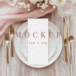 May include: A mockup of a place setting with a white plate, gold cutlery, and a white card. The card has the word "MOCKUP" printed on it. A pink cloth and flowers are in the background. The setting is on a light wood surface.