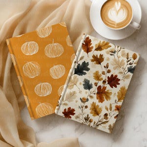 May include: Two hardcover journals with fall-themed designs. One journal has an orange cover with white pumpkin illustrations. The other journal features a cream-colored cover with autumn leaves in shades of brown, green, and gold. A cup of coffee sits nearby.