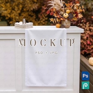 May include: A white rectangular table runner draped over a white wooden console table. The text MOCKUP and PSD + JPG are printed on the runner. A ceramic vase with autumn foliage and small pumpkins sits on the table next to a stack of folded napkins.