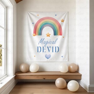 K&ouml;nnte beinhalten: Ein wei&szlig;es Banner mit einem bunten Regenbogen-Design, Sternen und dem Text "Magical DEVID" in Blau. Das Banner h&auml;ngt &uuml;ber einer Holzbank mit mehreren Ballons in neutralen Farben.