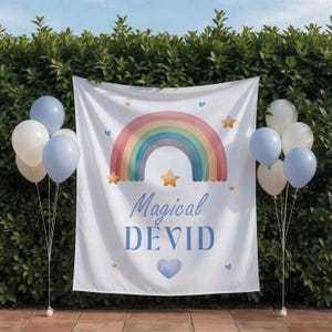 May include: A white banner with a rainbow design, stars, and the text "Magical DEVID" in blue. The banner is decorated with blue and white balloons. The background is a green hedge.