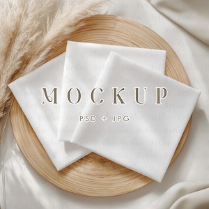 Puede incluir: Imagen de maqueta con tres servilletas cuadradas blancas dispuestas sobre un plato redondo de madera marrón claro. La palabra "MOCKUP" se muestra en el centro, con "PSD + JPG" debajo. Hierba de la pampa seca y tela blanca en el fondo.