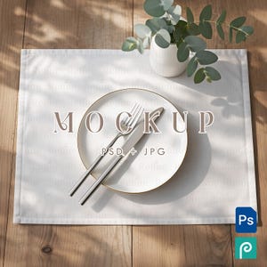 May include: A white placemat with a white plate featuring a gold rim, a fork, and a knife. A small white vase with green leaves sits in the background. The word "MOCKUP" is printed on the placemat.