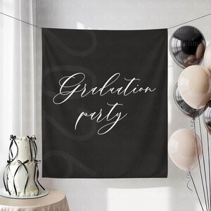 May include: A black backdrop with the words "Graduation party" in white script. A two-tiered white cake with black ribbon and balloons in black and beige are on the left and right sides of the backdrop.