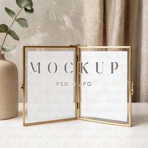 May include: A hinged, gold-colored metal photo frame with clear glass panels. The frame is open, displaying the word "MOCKUP" and the text "PSD + JPG" on the white background. The frame is on a white surface.