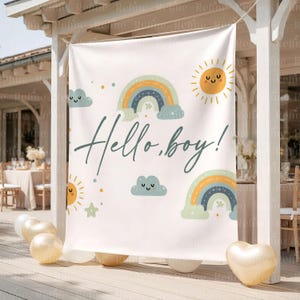 Op de afbeelding: Een cr&egrave;mekleurige banner met de tekst "Hello, boy!" in een scriptlettertype, versierd met regenbogen, lachende zonnen, wolken en sterren in pastelkleuren. Goudkleurige ballonnen staan onderaan. De banner wordt buiten getoond.