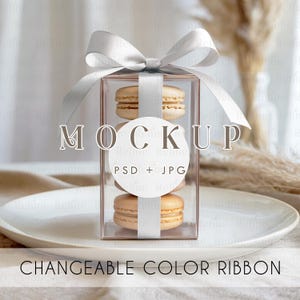 May include: A clear box tied with a silver ribbon, containing three macarons. The box is on a white plate. The text "MOCKUP" and "CHANGEABLE COLOR RIBBON" are visible.
