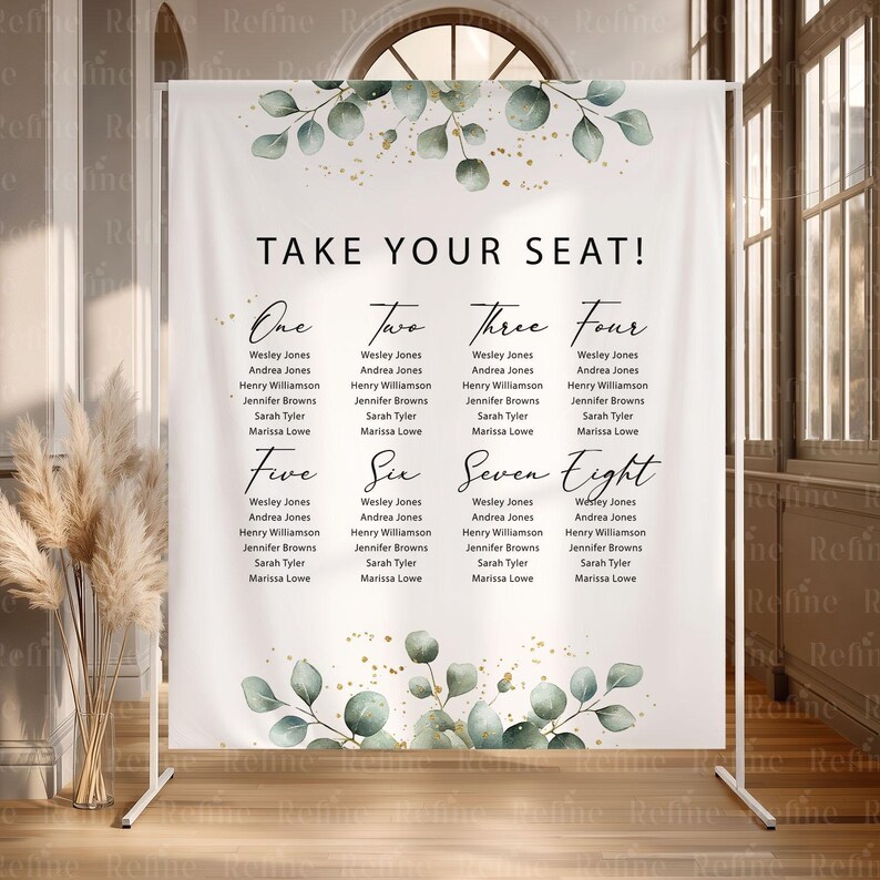 May include: A white wedding seating chart banner with the text "TAKE YOUR SEAT!" and guest names listed under numbers one through eight. The banner has a floral design with green leaves and gold accents, and is displayed on a white metal stand.