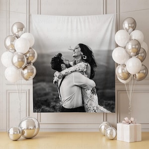 May include: A black and white tapestry featuring a couple embracing, surrounded by silver and white balloons. A gift box with a pink bow and disco balls complete the celebratory scene. The tapestry is hanging on a wall.