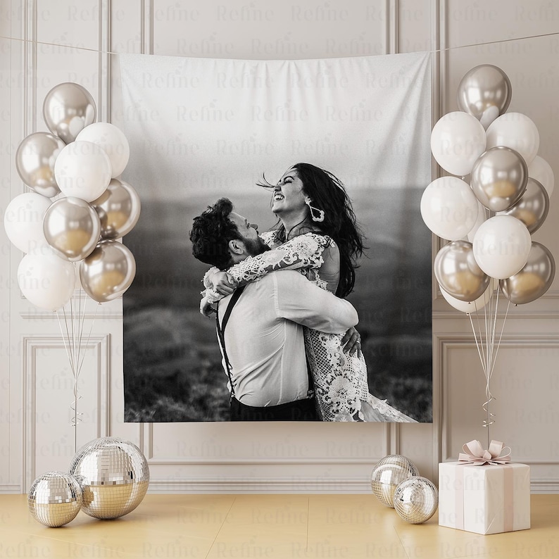 May include: A black and white tapestry featuring a couple embracing, surrounded by silver and white balloons. A gift box with a pink bow and disco balls complete the celebratory scene. The tapestry is hanging on a wall.