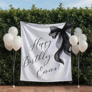 May include: A white banner with a black bow and the words "Happy Birthday Emma!" in elegant script. White balloons are on either side of the banner. The banner is hanging in front of a green hedge.