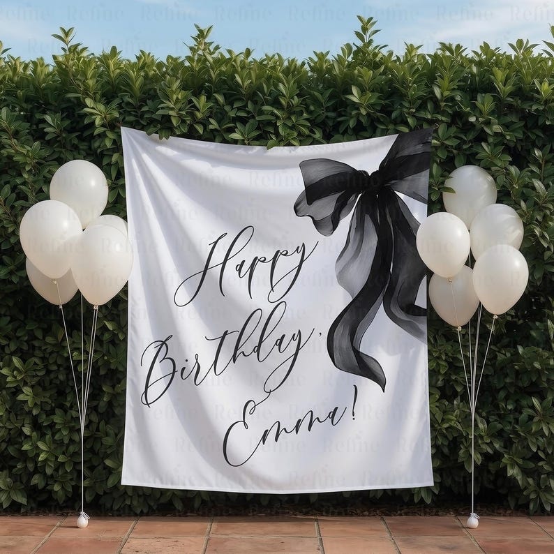 May include: A white banner with a black bow and the words "Happy Birthday Emma!" in elegant script. White balloons are on either side of the banner. The banner is hanging in front of a green hedge.