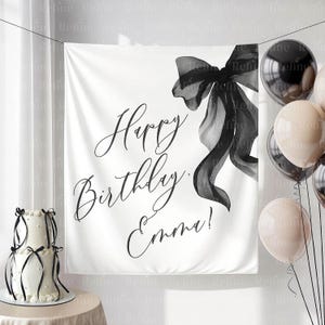 May include: A white banner with the text "Happy Birthday Emma!" in elegant script, accented by a black bow illustration. A two-tiered cake with black ribbon and balloons complete the birthday theme.