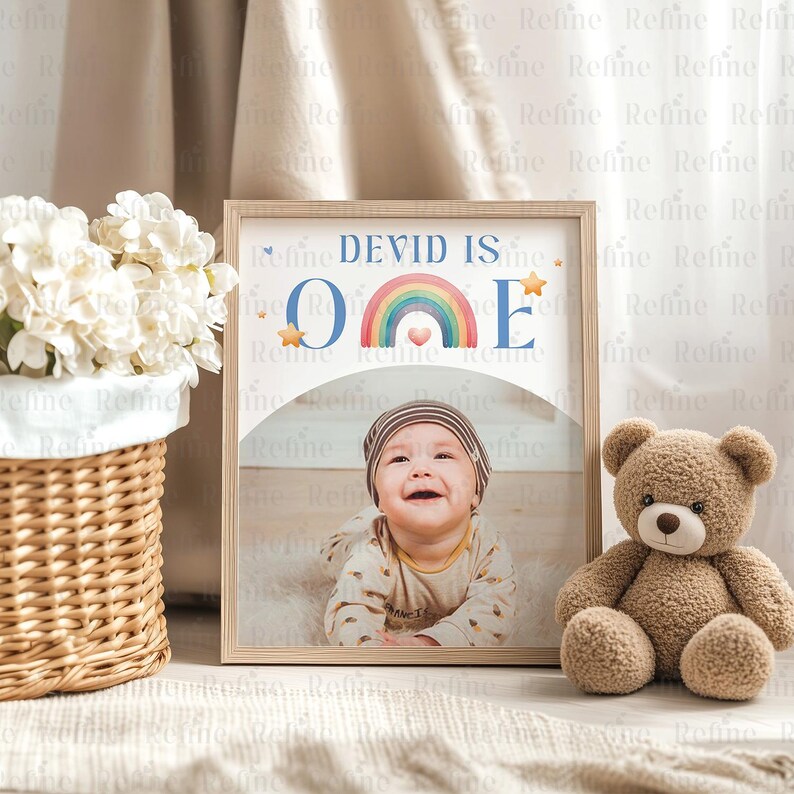 May include: A framed photo with a rainbow and the text "DEVID IS ONE" in blue letters. A baby boy is smiling at the camera in the photo. The frame is made of wood and has a light brown finish.