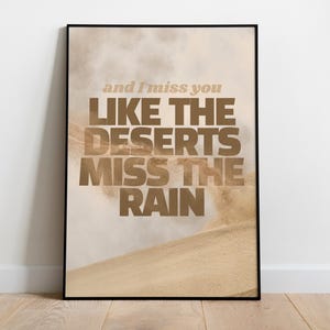 Puede incluir: Un cuadro enmarcado con un diseño de temática desértica. La obra presenta el texto "and I miss you LIKE THE DESERTS MISS THE RAIN" en una fuente marrón, sobre un fondo de dunas de arena y un cielo brumoso. La impresión está en un marco negro.