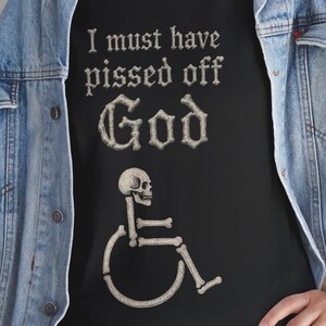 Puede incluir: Camiseta negra con la frase "I must have pissed off God" en fuente gótica. Debajo, una figura esquelética en silla de ruedas. Se usa con una chaqueta vaquera azul y vaqueros.